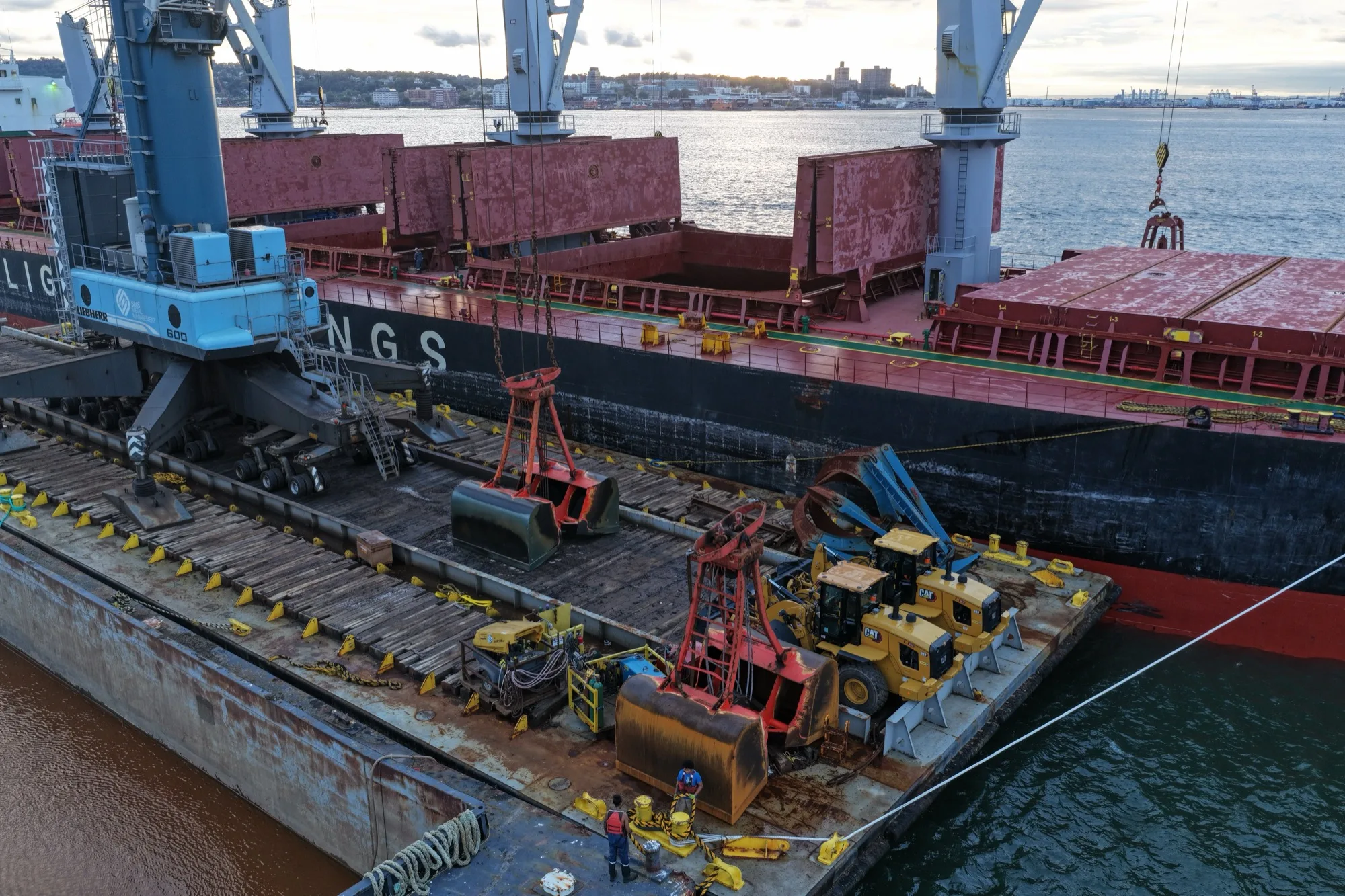 Grab buckets and wheel loaders on the crane barge alongside the ship, with a crew member at the mooring line