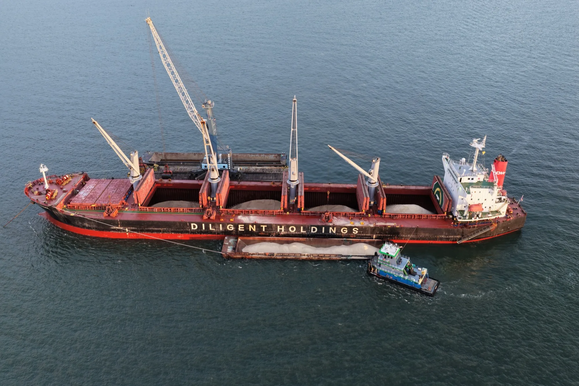 Aerial view of the Supramax vessel at anchor, holds open and full of rock salt, with a salt barge and tug alongside