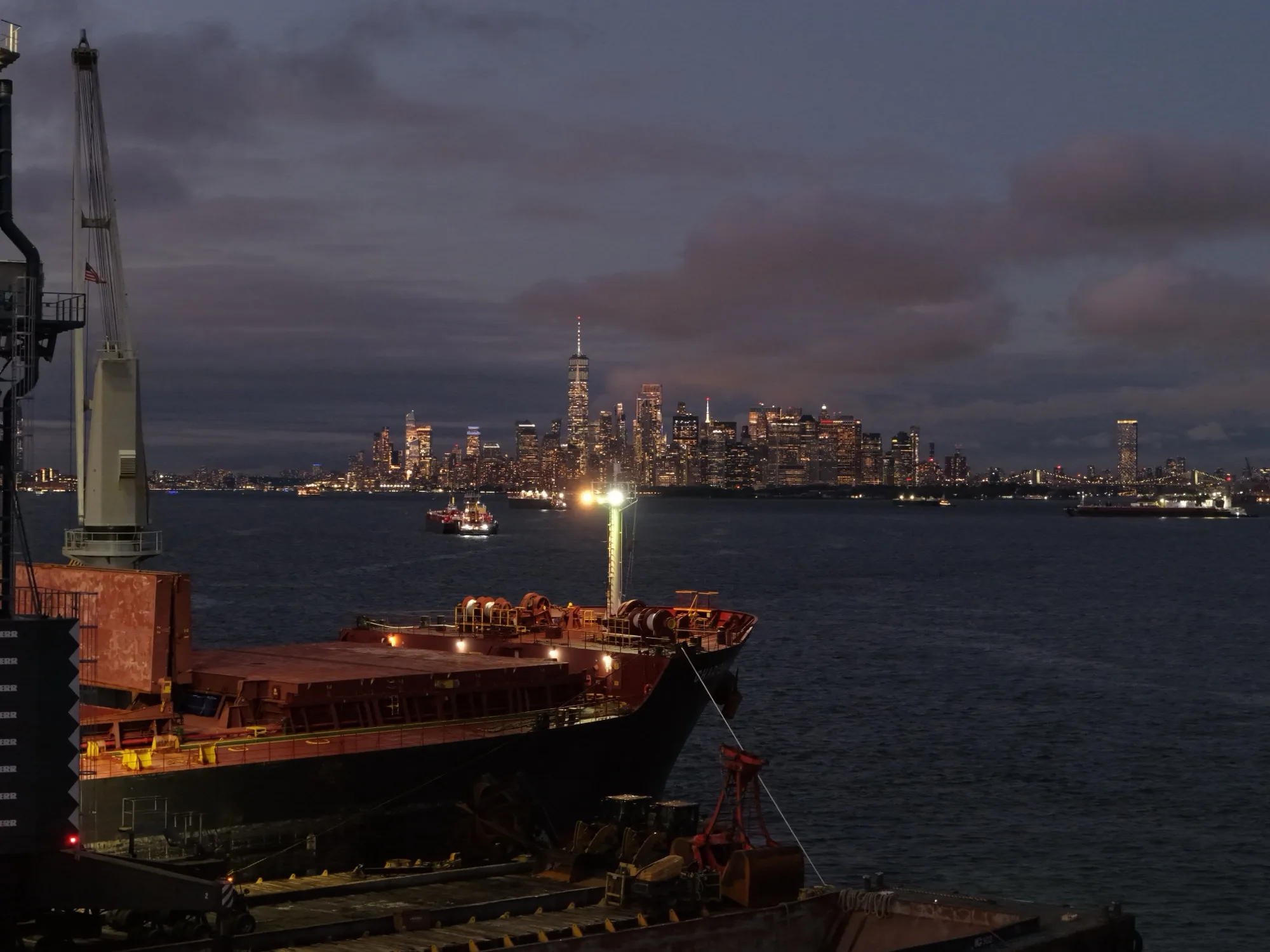 The bow of the ship at dusk, seen from the crane barge, with the lit Lower Manhattan skyline across the harbor
