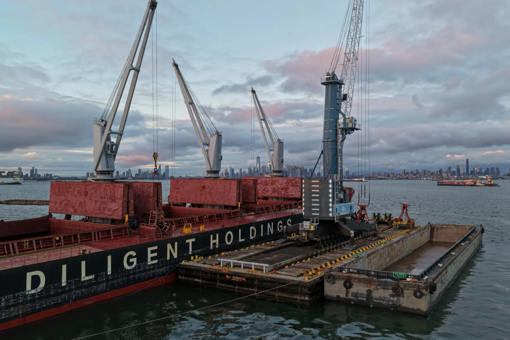 The ship's cranes and the crane barge alongside at dusk, the Manhattan skyline on the horizon