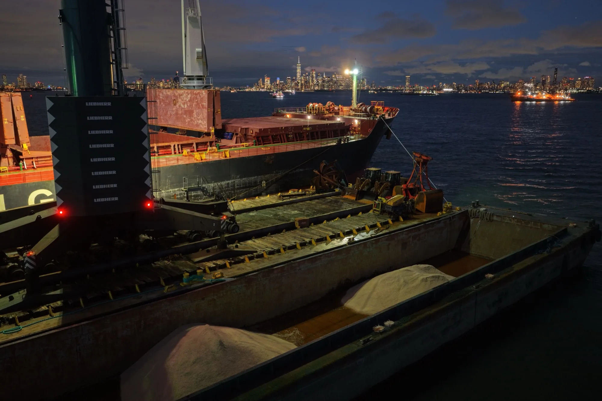 Discharging at night: the crane barge alongside the ship, rock salt piled in the barge and the Manhattan skyline in the distance