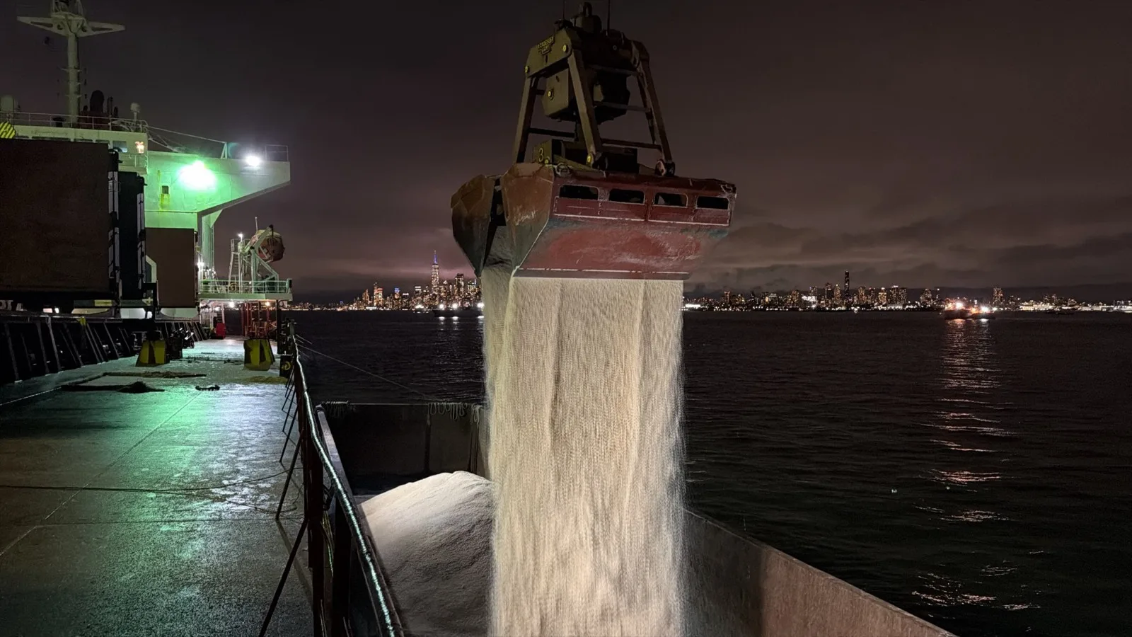 At night, golden rock salt pours from the grab into the barge, with the Manhattan skyline across the harbor