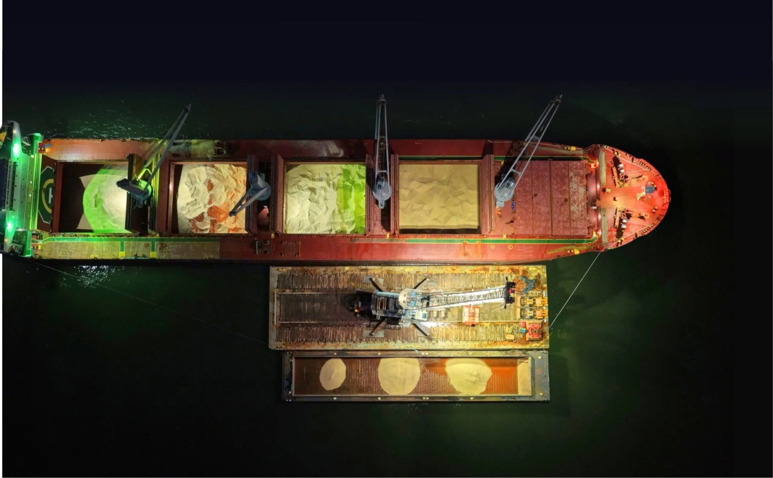 Overhead night view of the ship's open holds of rock salt, with the crane barge unloading into a salt barge alongside