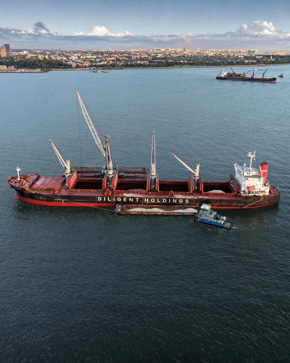 Aerial view of the ship at anchor off the harbor shore, a salt barge and a tug alongside
