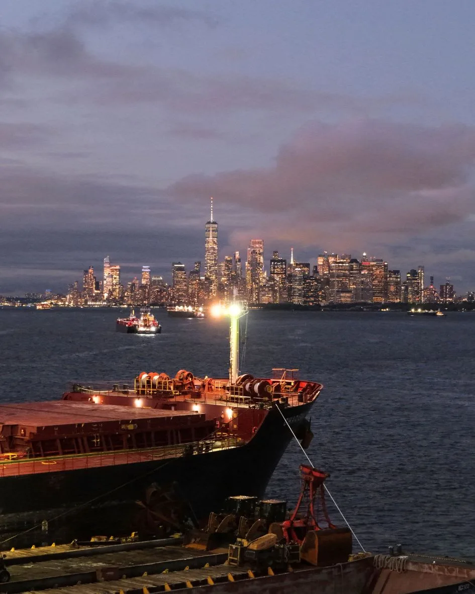 The ship's bow and the crane barge at dusk, the lit Lower Manhattan skyline across the harbor