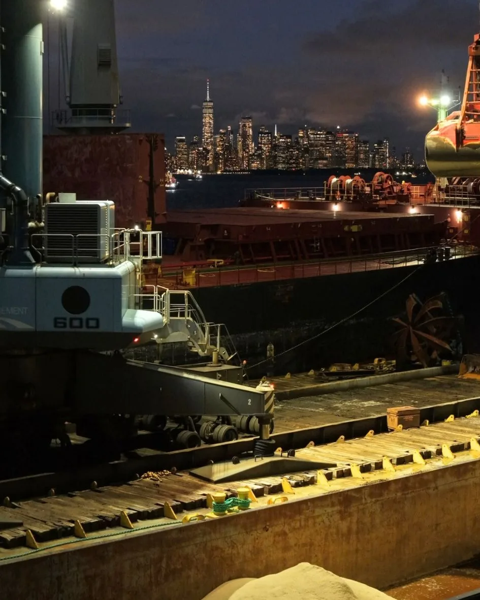 The crane barge beside the ship at night, the lit skyline across the harbor