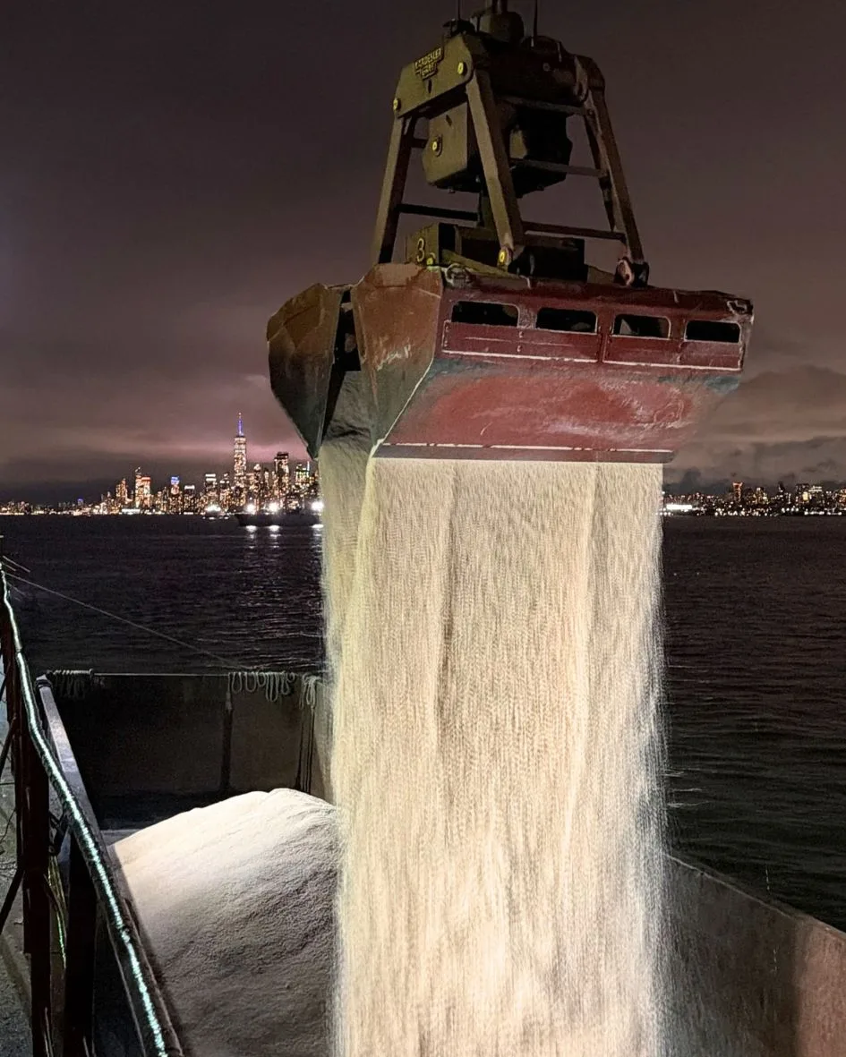 At night, golden rock salt pours from the grab into the barge, the Manhattan skyline across the water