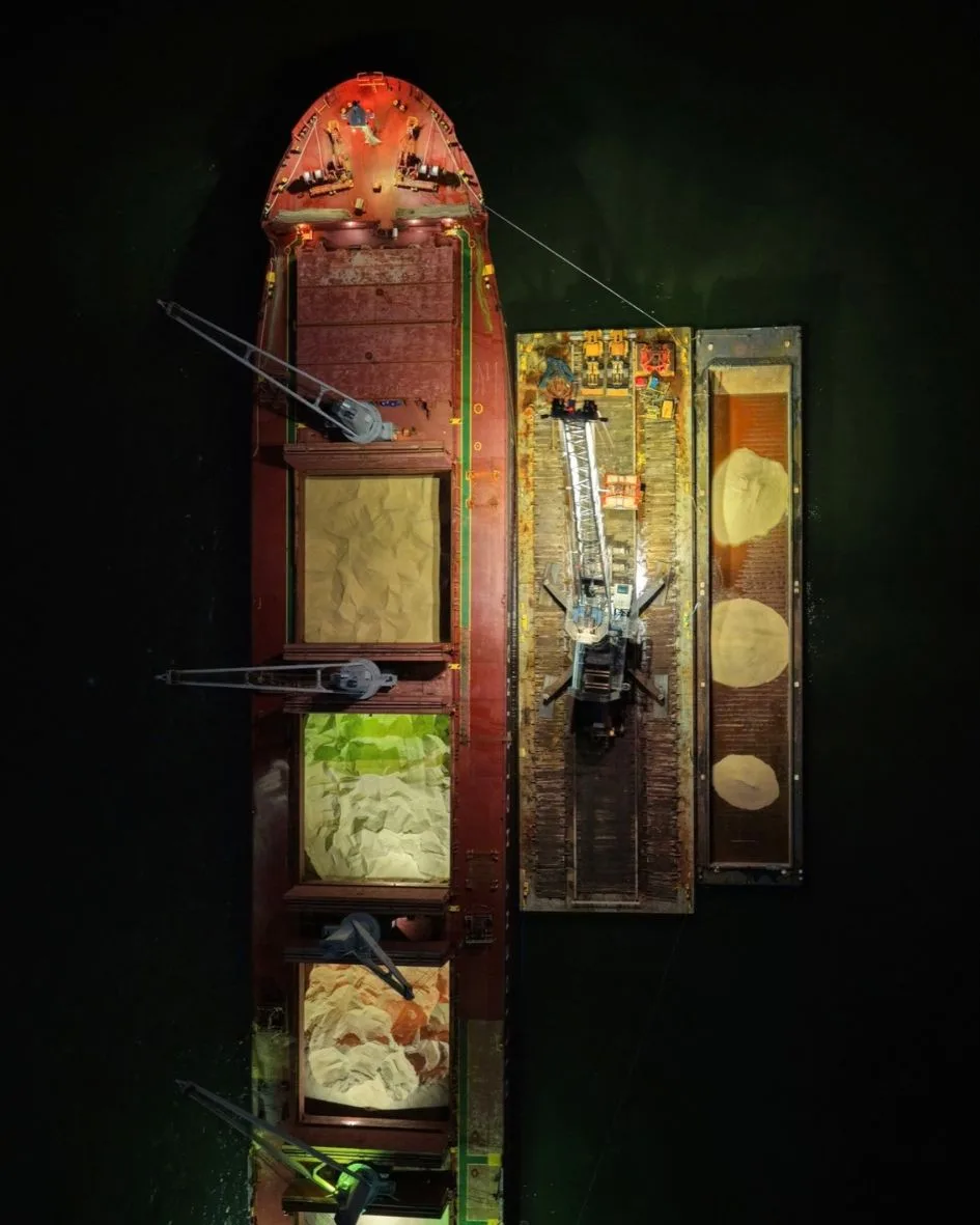Top-down night view of the ship's lit holds of rock salt, with the crane barge and a salt barge alongside