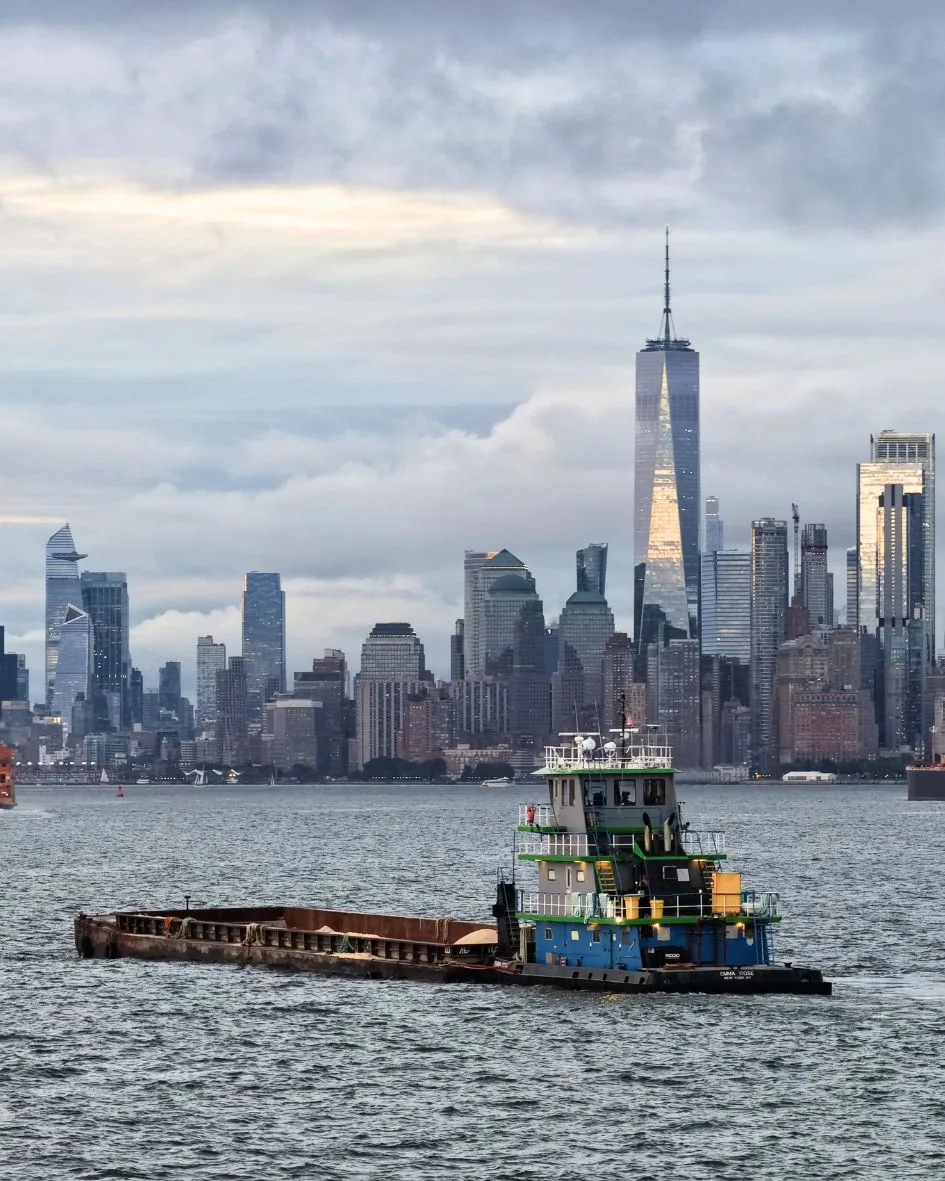 A tug pushing a barge of rock salt across New York Harbor, the Lower Manhattan skyline behind
